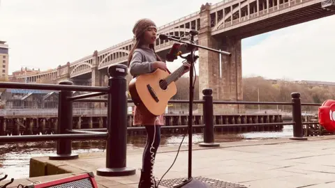 Reuben Abraham Nine-year-old Skye busking on Newcastle Quayside. She is wearing a beanie hat and is playing an acoustic guitar. She is singing into a microphone on a stand. The River Tyne and the High Level Bridge are in the background behind her.