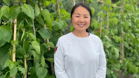 Lijia Ashcroft is standing in a garden smiling. Next to her are tall green bean plants. She has black hair pulled back from her face and is wearing a pale grey sweatshirt.
