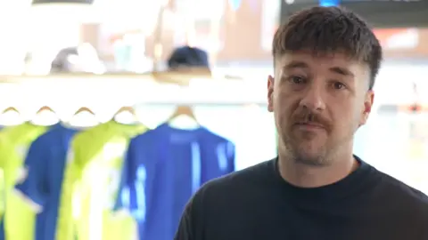 Jamie Niblock/BBC James Cole with short hair and a dark top. In the background are shirts hanging in a window.