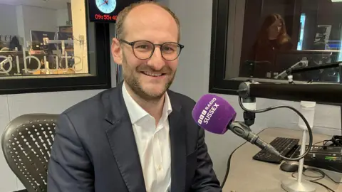 A man wearing glasses, wearing a black suit and white shirt, is sitting inside a BBC radio studio. 