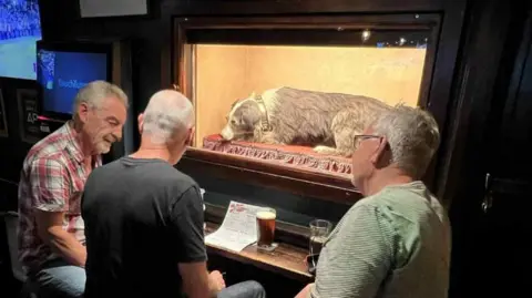 Nigel Meadows Three men are sitting at a small table mounted on the wall, and drinking pints. In front of them is the taxidermised dog, Wandering Willie, who has grey and white fur. Wille is behind a glass panel on a red cushion.