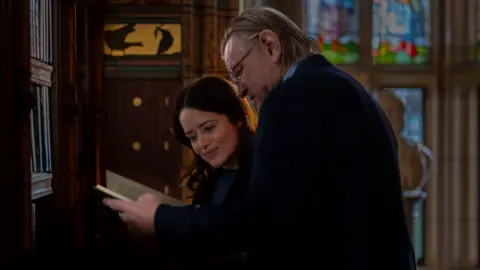 Roadside Attractions A still taken from the H is for Hawk film. Actors Claire Foy and Brendan Gleeson are standing next to each other while looking into the pages of a book in what looks like a panelled room with stained glass windows.