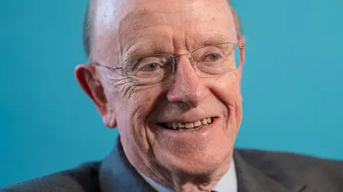 SOSE A smiling older man with glasses and a receding hairline against a blue background