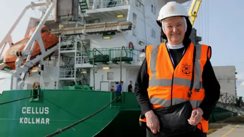 BBC/Ed Hanson Sister Mary Scholastica stands wearing a high vis orange jacket in front of a green merchant vessel at the Port of Sunderland in 2017. 