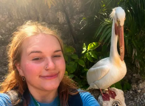 Amy is smiling at the camera, she has ginger hair and brown eyes and nose piercings, she is wearing a blue t shirt. On her left is a large white bird with an orange beak. 