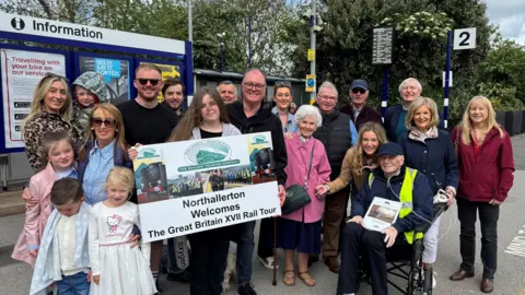 An elderly man in a wheelchair with his family. About 20 people including children. They are stood on a railway platform holding a sign that reads 'Northallerton Welcomes The Great Britain XVII Rail Tour'.