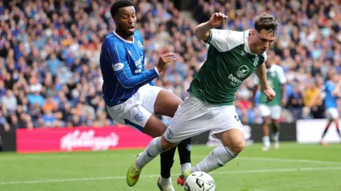 PA Media Two football players mid tussle. One is wearing blue and the other green. A football is visible in front of them and a stand of fans behind.