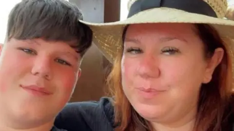 Family photo Morgan and Victoria Hopkins pictured sitting close together. Morgan wears a black t-shirt and has short brown hair and Victoria wears a black top with a v-neck and has red bobbed hair under a straw hat
