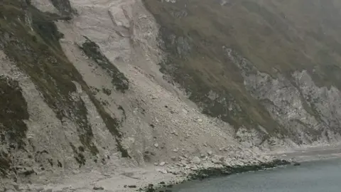 White rockfall and large boulders pour down from grassy cliffs to the edge of the sea