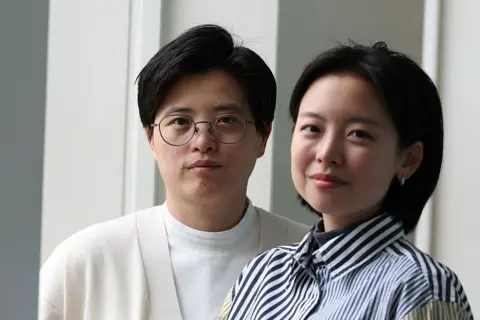 Adrian Dennis / AFP via Getty Images Yang Shuang-zi has short dark hair, wears thin, black rimmed glasses and wears a cream cardigan over a white t-shirt. Standing next to her, Lin King has slightly longer, shoulder length dark hair, and wears a blue and white striped shirt.