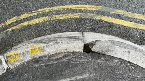 Close-up of part of the kerb from the top. It is scratched and scraped with a rough chunk taken from the top. Scuff marks on the yellow lines of the road.