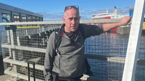 A bald middle-aged man standing with his hands against a wire fence at a ferry port, with a large vessel in the background. He is wearing a grey half-zip fleece, black t-shirt and has a pair of black rimmed glasses balancing on his head,