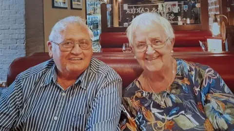William and Marlene Mumford sit smiling in a diner-style restaurant, on a red leather seat. They both have short white hair and glasses. William is wearing a blue and white striped shirt, and Marlene is wearing a blue floral top. 