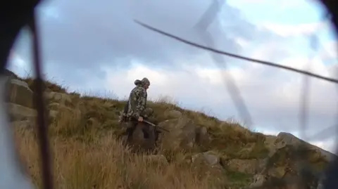 RSPB secret filming A man with a hood up on his head dressed in camouflage gear with a shotgun in his hands is walking across moorland. He is walking along some rocks and grassy land. Some black lines are visible in this screen grab from the covert footage and they are probably some grass in front of this covert camera set up for surveillance. 