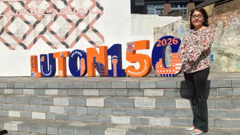 Amy Holmes/BBC Tahmina Saleem stands on steps beside large "LUTON 150" display letters in a sunny outdoor setting.