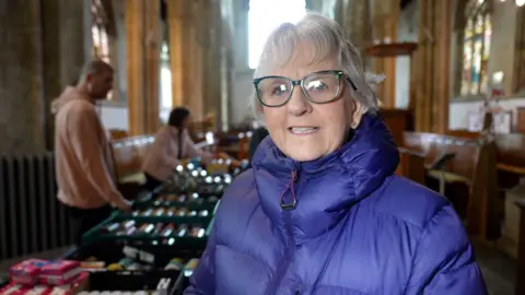 A woman stands inside a church, facing the camera, wearing glasses and a padded purple jacket. Behind her are helpers and prduce for St Augustine's food bank. There are stained‑glass windows, stone columns and wooden pews visible in the background. 