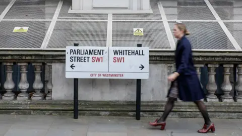 A pedestrian passes a street sign display the start of Parliament Street, left, and Whitehall, right, in the Westminster district of London, UK.