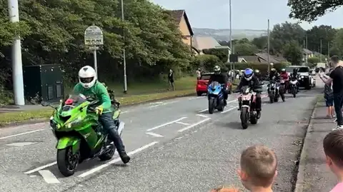 Family photograph A number of motorbikes in a special vehicle parade for a young boy and girl in Bury. Young children are seen among the people lining the street for the parade. It is a dry day. Hills can be seen in the background.