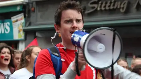 Conradh na Gaeilge Pádraig Ó Tiarnaigh has short brown hair and is wearing a red t-shirt and blue backpack. He is speaking into a microphone and has a clear/white earpiece.