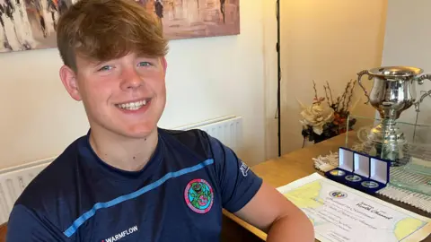 Oscar has brown hair pushed forward, he is smiling. He is wearing a navy blue T shirt with a light blue strip across the chest. He is sat by a table, on which is a certificate, three medals in blue cases and a large silver trophy. 