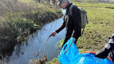 Wear Rivers Trust A volunteer from Wear Rivers Trust stands by a stream and is picking up waste with a litter-picker. They are holding a bright blue bin bag. The volunteer has short grey hair and is wearing all black outdoor clothing, a grey cap and a grey rucksack.