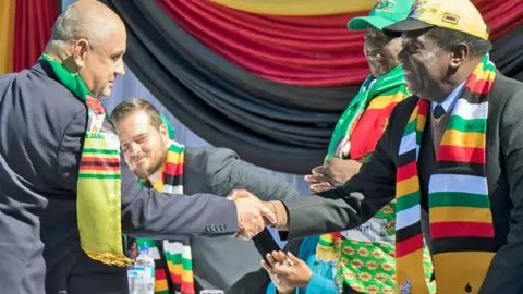 AFP via Getty Images President Emmerson Mnangagwa in a yellow cap and scarf in the Zimbabwean flag's colours shakes the hand of a white man wearing a yellow Zanu-PF scarf at a political campaign meeting in July 2018 for members of the white farming and business community.