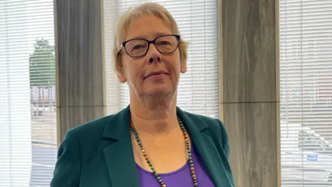 A woman with short blonde hair and wearing glasses, a green jacket, a purple top and a beaded necklace, stands next to an office window.