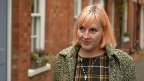 Sam Read/BBC Jennifer Wilson-Marklew with light-coloured hair stands on a residential street lined with red-brick houses. The person is wearing a patterned coat over a checked top and a pendant necklace. The background is softly blurred, showing house windows and a narrow pathway between the buildings.