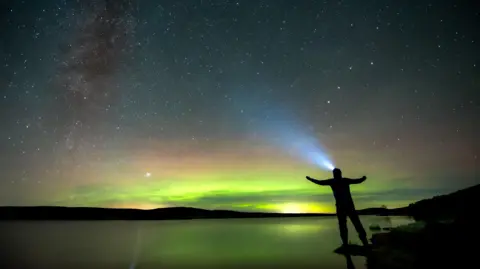 Alan Rankin The silhouette of a man wearing a head torch in front of the green hues of the northern lights.