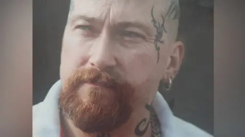 Ian Michael A close-up image of a man with a brown beard and tattoos on the left and right of his face. He is looking to his right on the photo.