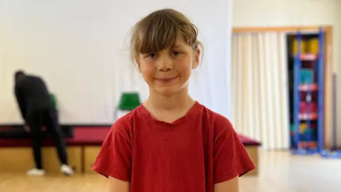 A girl in a red shirt looking at the camera and smiling.
