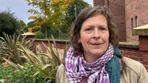 A woman with shoulder-length brown hair wears a beige jacket and a purple scarf and stands in front leafy foliage and a red-brick property.