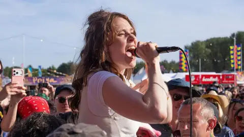 Getty Images Kate Nash pictured in a white top singing into a handheld microphone while standing among a large crowd at an outdoor music festival. Audience members surround the performer, with some filming on smartphones. Colourful festival banners, food stalls and a line of trees are visible in the background under a bright blue sky.
