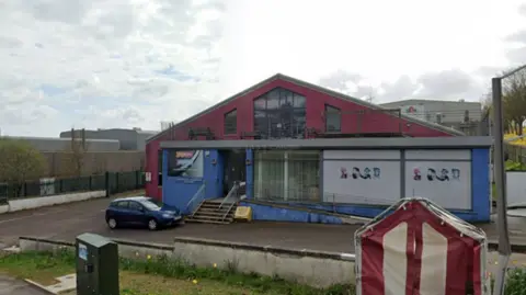 Aberdeen's Northsound building. It has a set of stairs to the front and is over two floors. The first floor is painted blue and has large windows. The upper floor has a balcony with benches and large glass doors and windows. It is painted red. A blue car is parked outside the main door. 