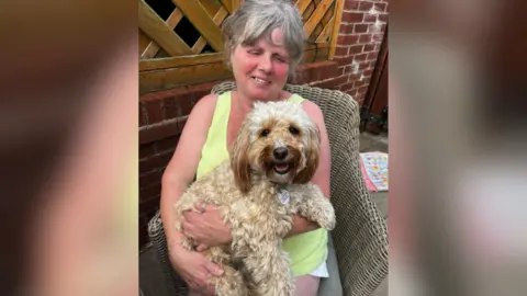 Woman with greying hair proudly holds dog - a blonde Cavapoo. The pair sit outside in a wicker chair. 