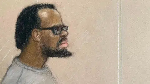 Court sketch of Simon Levy, wearing a long sleeved grey top and thick rimmed glasses. He has short dark hair which appears to be receding, and a beard.