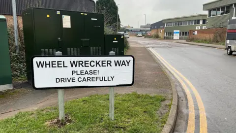 A road sign which has been covered up with the words "Wheel Wrecker Way, Please! Drive Carefully". A road with potholes is in the background with an industrial estate beyond. The first section of the road is maintained with no potholes.