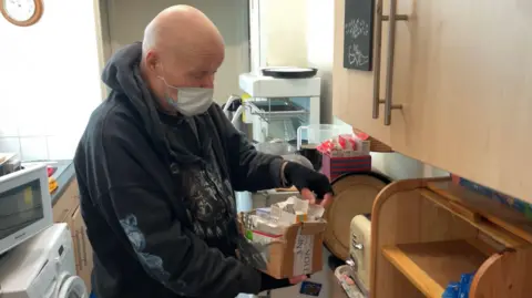 A man wearing a black hoodie and a surgical face mask sorts through boxes of medication on a kitchen worktop. He is reaching into a cardboard box filled with blister packs and medicine packets. Behind him are a microwave, a washing machine and other kitchen appliances.
