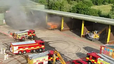 A number of fire engines on a concrete surface with thick smoke hanging in the air.