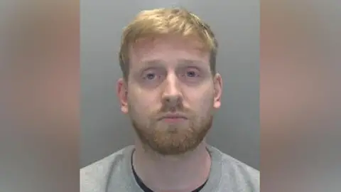 A police mugshot of Jakob Ireson. He has short ginger hair and beard, and is standing in front of a grey wall. He is wearing a grey jumper with a black T-shirt underneath.