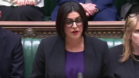 Mahmood wears a black jacket over a royal purple top. She wears her black hair in a chin length bob, with spectacles and red lipstick.