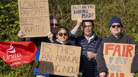 Four people hold signs which read "Lowest pay offer, best tap water" and "3.4%? Are you barking mad?" and "Is this an annual thing now?" and "Fair pay for fair work". A red Unite the union flag waves beside them