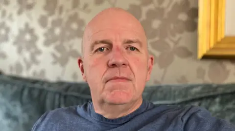 Lewis Adams/BBC Paul Maleary has a shaved head and is wearing a navy crew neck jumper. He is sitting on a blue sofa and looks serious.