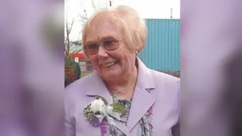 A woman with short white hair wearing dark glasses and a purple floral jacket.