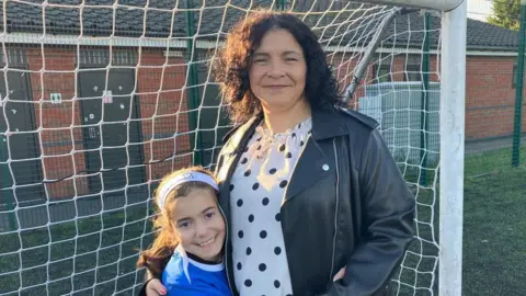 A woman with dark curly hair, black and white polka dot top and black leather jacket is smiling and hugging her daughter, who is dressed in a blue football kit and they are standing in front of a goal on a football pitch.