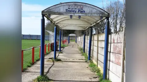 A Tesco trolley shelter is pictured in a football ground with goalposts and green grass in the background