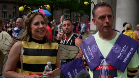 A woman with long brown hair and wearing a bee outfit is smiling. She is standing next to a man in a light green top and both are holding purple flags with 'Manchester Hospital School' and 'Sponsor us' written on them. 
