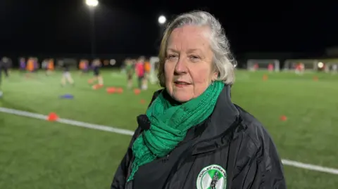Yvonne stands smiling at a training session wearing her black Tunley Athletic-branded coat and green scarf