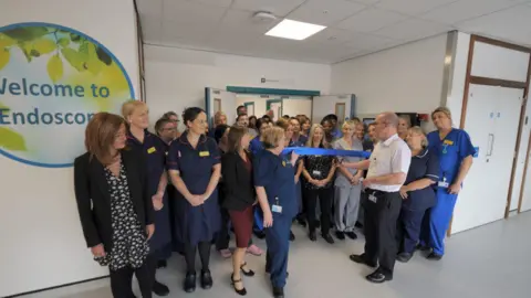 A crowd of hospital staff stand in front of open double doors. On the wall next to them is a sign that reads "Welcome to Endoscopy", in blue writing. Staff are holding a blue ribbons, and a man is holding scissors up to cut it in half