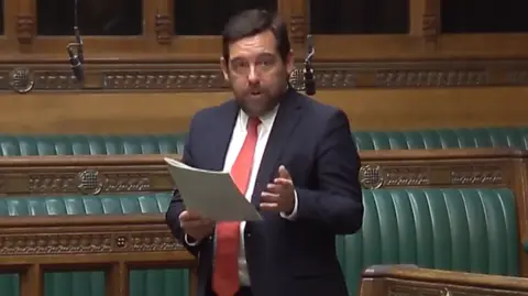 David Baines is standing in parliament. He has short brown hair, and is wearing a red tie, white shirt, and navy jacket. He is speaking, and reading from a sheet of paper.